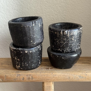 May include: Four black, textured, cylindrical bowls with a rustic appearance are stacked on a wooden surface. The bowls have a dark, matte finish with lighter, distressed accents, suggesting a handcrafted or aged aesthetic. The bowls are arranged in two stacks.