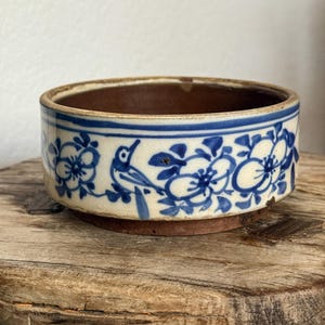 May include: A round ceramic bowl with a cream-colored base and a dark brown interior. The exterior features a hand-painted blue floral pattern interspersed with a small bird motif, framed by thin blue horizontal lines along the top rim.