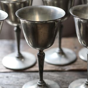 May include: A collection of silver-toned metal goblets featuring a smooth, rounded bowl, a slender stem with a subtle decorative node, and a wide, flat circular base. The metallic surface displays a soft, reflective sheen with visible texture.