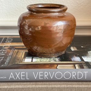 May include: A small, round, brown ceramic pot with a glossy, uneven glaze and a wide, thick rim. The pot features a bulbous body with subtle indentations along the surface, resting on a flat base with visible signs of wear and texture.