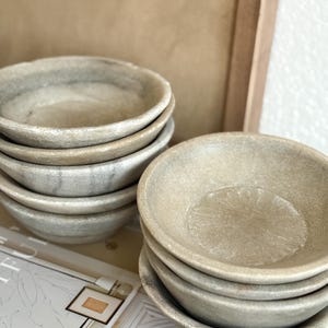 May include: A stack of small, round, stone-like bowls in varying shades of gray and beige. The bowls have a textured surface and a shallow, dish-like shape. The bowls are stacked in two separate piles, suggesting a set for serving or display.