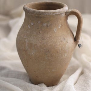 May include: A rustic, tan-coloured pitcher with a handle. The earthenware jug has a rounded body and a wide opening, with a slightly textured surface. The pitcher sits on a white, textured fabric.