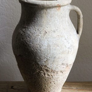 Puede incluir: Un jarrón de cerámica de color claro y desgastado, con asa. El jarrón tiene una boca ancha y una superficie texturizada, mostrando signos de edad y desgaste. Se encuentra sobre una superficie de madera, sugiriendo una estética rústica o antigua.