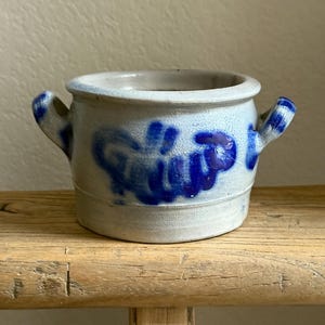 Puede incluir: Una pequeña olla redonda de gres con dos asas. La olla es de color azul claro con letras y detalles en azul oscuro. La olla está sobre una superficie de madera.