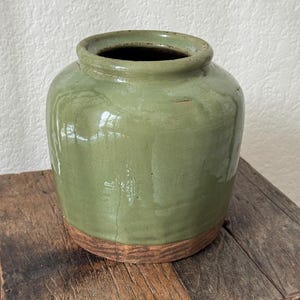 May include: A sage green ceramic jar with a wide, rounded body and a short, thick neck. The upper portion features a smooth, glossy glaze, while the base remains unglazed, revealing a textured, natural brown clay surface with visible cracks.