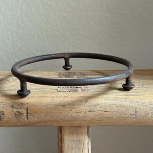 May include: A circular, dark brown metal plant stand with three short legs. The stand is made of wrought iron and sits on a light brown wooden surface. The stand is designed to hold a small potted plant.