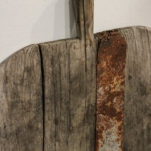 May include: Close-up of a rustic wooden cutting board with a handle. The board is made of weathered wood with visible grain and cracks. A rusty metal strip reinforces the board. A small nail is at the top of the handle.