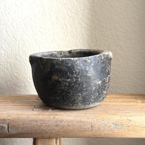 May include: A small, dark gray, stone bowl with a rough, textured surface. The bowl is round with a slightly irregular rim and sits on a weathered wooden bench. The background is a light beige wall.