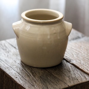 May include: A cream-colored ceramic crock with a wide circular opening, a smooth glazed finish, and two small, triangular-shaped handles protruding from opposite sides of the upper body. The vessel features subtle horizontal ridges near the rim.