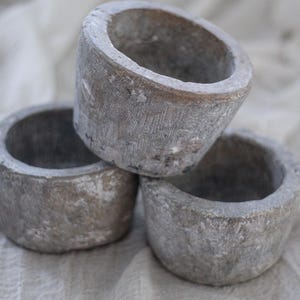 May include: Three stacked, weathered stone ring bowls. The bowls are a muted gray color with textured surfaces and a rough, aged appearance. They sit on a light-colored, textured fabric.