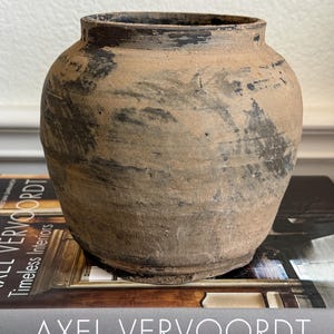 May include: A round, wide-mouthed ceramic vessel with a textured, matte finish in shades of tan and brown. The surface features irregular, dark charcoal-colored markings and weathered patches, giving it a rustic, aged appearance with a wide rim.