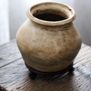Pode incluir: Um vaso de cerâmica bege de formato arredondado com uma abertura larga e uma superfície texturizada e desgastada. O recipiente apresenta uma borda espessa e arredondada, apoiado sobre um suporte metálico circular escuro com pés pequenos.
