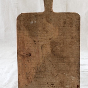 Puede incluir: Una tabla de cortar rectangular de madera con un asa. La tabla es de madera marrón claro con vetas visibles y algunas pequeñas imperfecciones. El asa es una extensión estrecha y rectangular de la tabla.