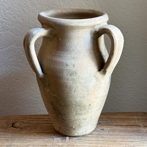 May include: A beige, weathered clay vessel featuring a wide, circular opening and two symmetrical side handles. The surface displays a matte, textured finish with subtle variations in tone, showing signs of age and organic, earthy wear.