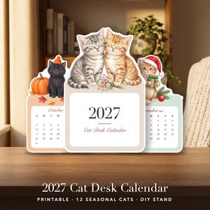 May include: A 2027 cat desk calendar featuring seasonal illustrations of kittens. The center panel shows two kittens with flowers, while side panels display October with a black cat and pumpkin, and December with a kitten in a Santa hat.