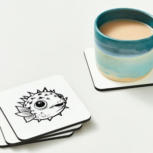 May include: Four white coasters with black line art of a pufferfish on the top coaster. The coasters are stacked on top of each other.