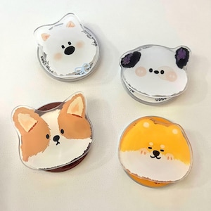 May include: Four clear acrylic phone grips featuring cartoon dog faces. The grips include a white dog with a bow, a white dog with black spots, a corgi, and a yellow dog. Each grip has a circular base.