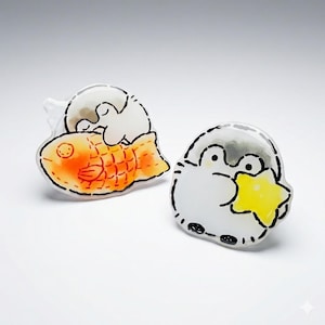 May include: Two whimsical, cartoon-style pins. One features a white penguin on an orange fish-shaped pastry. The other pin shows a penguin holding a yellow star. Both pins have black outlines and are set against a white background.
