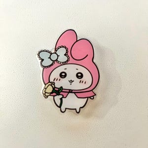 May include: A cartoon-style character with a pink hood and a light blue bow. The character is holding a yellow flower. The design has a white body and a pink cape. The image is on a white background.