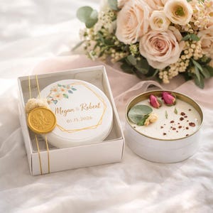 May include: A white candle in a gold-rimmed tin, decorated with dried flowers and petals. Next to it, a white round box with a floral design and the text "Megan & Robert 01.15.2026" is tied with gold string and a wax seal. A bouquet of roses is in the background.