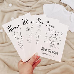 May include: A hand holds four white coloring cards with black outlines of letters and illustrations. The cards read "B is for", "D is for", "F is for", and "I is for Ice Cream". A white baby outfit and hat are in the background.