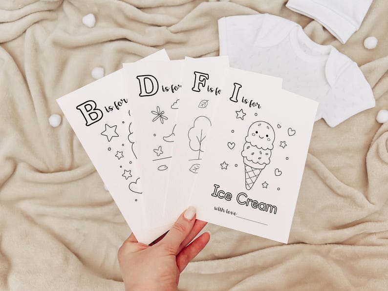 May include: A hand holds four white coloring cards with black outlines of letters and illustrations. The cards read "B is for", "D is for", "F is for", and "I is for Ice Cream". A white baby outfit and hat are in the background.