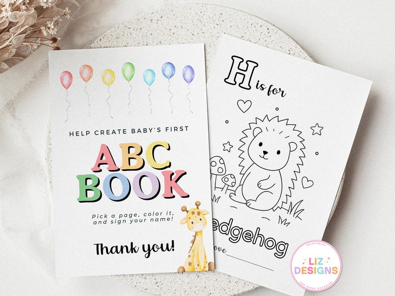 May include: Two white pages of a baby ABC book. One page has colorful balloons and the text "Help Create Baby's First ABC Book". The other page has a hedgehog illustration and the text "H is for hedgehog". A giraffe illustration is also present.