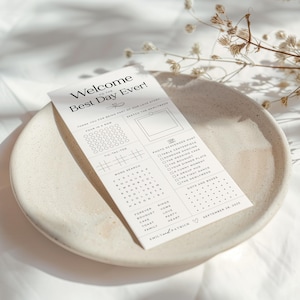 May include: A white wedding activity card with the text "Welcome to the Best Day Ever!" sits on a beige ceramic plate. The card includes a word search, tic-tac-toe, and a photo scavenger hunt. Dried flowers are in the background.