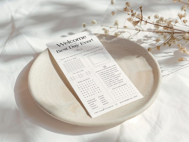 May include: A white wedding activity card with the text "Welcome to the Best Day Ever!" sits on a beige ceramic plate. The card includes a word search, tic-tac-toe, and a photo scavenger hunt. Dried flowers are in the background.