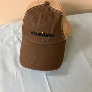 May include: A brown baseball cap with a mesh back and a curved brim. The front of the cap features the text "etrailer.com" in black. The cap has a distressed look.