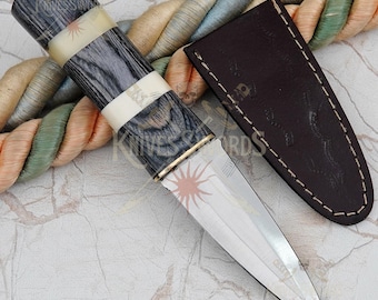 May include: A dagger with a polished silver blade and a handle featuring alternating bands of dark gray wood and cream-colored material. A brown leather sheath is next to the dagger. The handle has a brass-colored ring at the base of the blade.