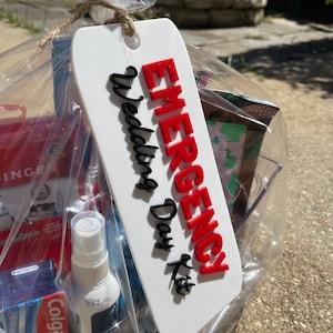 Pode incluir: Uma cesta de presente embrulhada em celofane transparente com uma etiqueta branca que diz "EMERGENCY Wedding Day Kit" em letras vermelhas e pretas. A cesta contém vários itens, incluindo um borrifador, pasta de dente e um pequeno kit de costura.