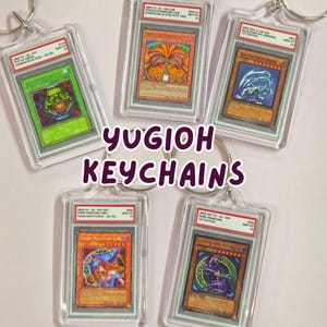 Pode incluir: Chaveiros Yu-Gi-Oh! com cartas colecionáveis, incluindo Dark Magician Girl e Blue-Eyes White Dragon. Cada carta está em um estojo protetor transparente com um porta-chaves prateado. O texto "YUGIOH KEYCHAINS" é exibido no centro.