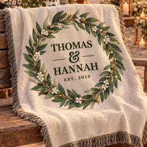 May include: A cream-colored woven throw blanket featuring a decorative olive branch wreath with green leaves and white flowers. The center text reads THOMAS & HANNAH EST. 2019 in dark green lettering. The blanket has a fringed edge on all sides.