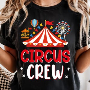 Puede incluir: Camiseta negra con un diseño de circo colorido. El diseño incluye una carpa a rayas rojas y blancas, una noria, un tiovivo y las palabras "CIRCUS CREW" en rojo y blanco. También aparece un globo aerostático.