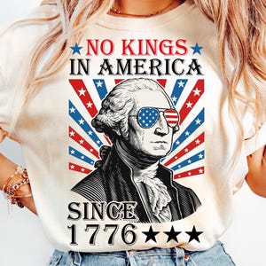 May include: Cream-colored t-shirt featuring a graphic of George Washington wearing sunglasses with a patriotic design. The shirt has the text "NO KINGS IN AMERICA" and "SINCE 1776" with stars. The design incorporates red, white, and blue colors.