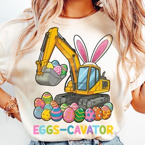 May include: A white t-shirt with a yellow cartoon excavator with bunny ears holding colorful Easter eggs. The text "EGGS-CAVATOR" is printed below the excavator.