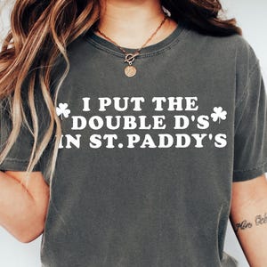 May include: Dark gray t-shirt with the text "I PUT THE DOUBLE D'S IN ST. PADDY'S" in white, with shamrock graphics. The shirt is a crew neck and has short sleeves.