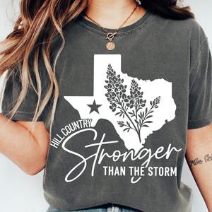 May include: Dark grey t-shirt with a white graphic of the Texas state outline, a star, and a floral design. The text reads "Hill Country Stronger Than The Storm". The shirt is a casual style, suitable for everyday wear.