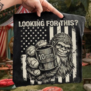 Könnte beinhalten: Schwarzes T-Shirt mit einem Grafikdesign, das ein Bigfoot-ähnliches Wesen zeigt, das einen 10-mm-Steckschlüssel hält. Das Design enthält ein amerikanisches Flaggenmotiv mit dem Text "LOOKING FOR THIS?" in verblichener Schrift. Das Bild ist in Schwarzweiß.
