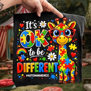 以下が含まれることがあります： 赤いメガネをかけた、パズルのピースで作られたカラフルなキリンのグラフィックが描かれた黒いクッション。「It's OK to be DIFFERENT」の文字と#AUTISMAWARENESSのハッシュタグ。デザインにはハート、花、虹が含まれています。