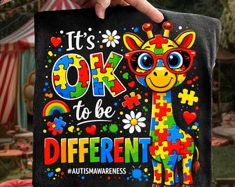 May include: Black pillow with a colorful graphic of a giraffe wearing red glasses, made of puzzle pieces. Text reads "It's OK to be DIFFERENT" with the hashtag #AUTISMAWARENESS. The design includes hearts, flowers, and a rainbow.