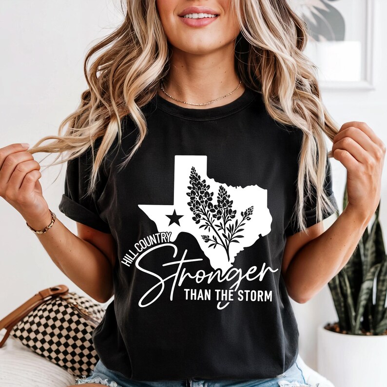 May include: Black t-shirt with a white graphic of the Texas state outline, a star, and a floral design. The shirt features the text "Hill Country" and "Stronger Than The Storm" in a stylish script.