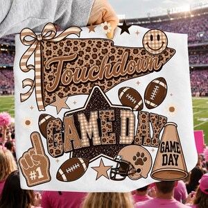 May include: A white t-shirt featuring a brown and tan graphic design with a leopard print pennant that says Touchdown, a large star with the text GAME DAY, footballs, a helmet, a megaphone, a foam finger, and a checkered smiley face.