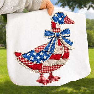 May include: White tote bag featuring a patriotic goose design. The goose is adorned with red, white, and blue patchwork, including stars and stripes, and a blue bow. The design is reminiscent of the American flag.