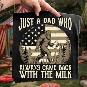 Puede incluir: Camiseta negra con un gráfico que presenta una criatura parecida a Bigfoot llevando leche, sobre un diseño de bandera estadounidense. El texto dice "JUST A DAD WHO ALWAYS CAME BACK WITH THE MILK."