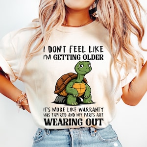 May include: Cream-colored t-shirt with the text "I DON'T FEEL LIKE I'M GETTING OLDER" above a cartoon turtle. The turtle is green with a brown shell. Below the turtle is the text "IT'S MORE LIKE WARRANTY HAS EXPIRED AND MY PARTS ARE WEARING OUT."