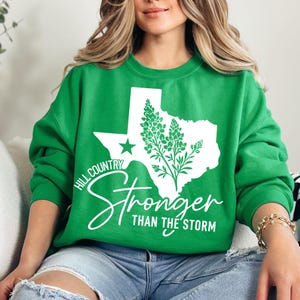 May include: A green crew neck sweatshirt with a white outline of Texas, a star, and a floral design. The text "Hill Country Stronger Than The Storm" is printed in white script. The sweatshirt is a casual style.
