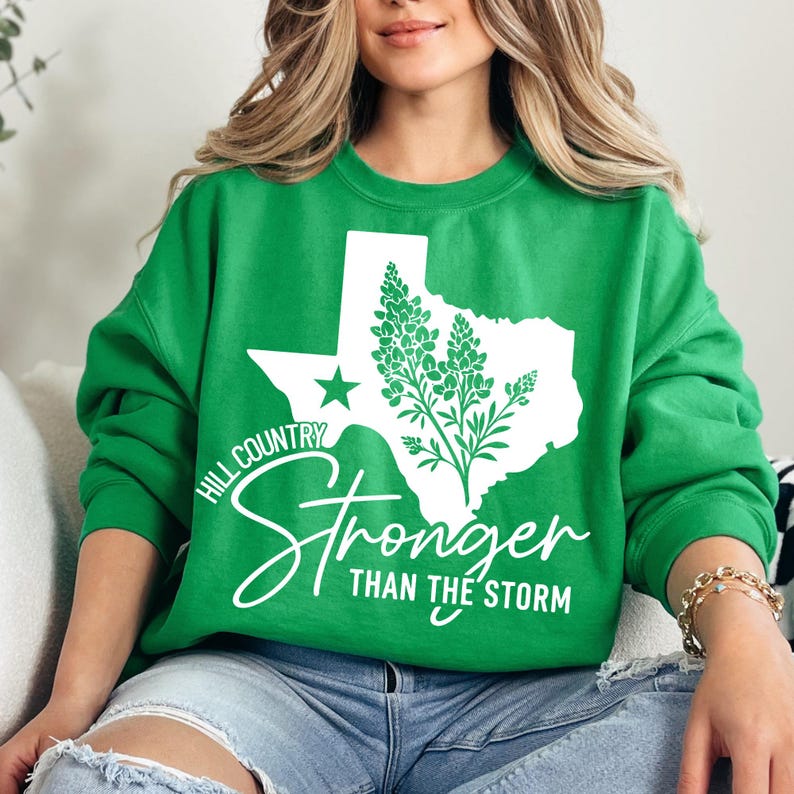 May include: A green crew neck sweatshirt with a white outline of Texas, a star, and a floral design. The text "Hill Country Stronger Than The Storm" is printed in white script. The sweatshirt is a casual style.