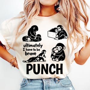 Puede incluir: Camiseta color crema con cuatro ilustraciones de monos negros y el texto "Ultimately I have to be brave PUNCH." El diseño incluye monos en varias poses, como abrazándose y jugando. La camiseta está hecha de un material suave.